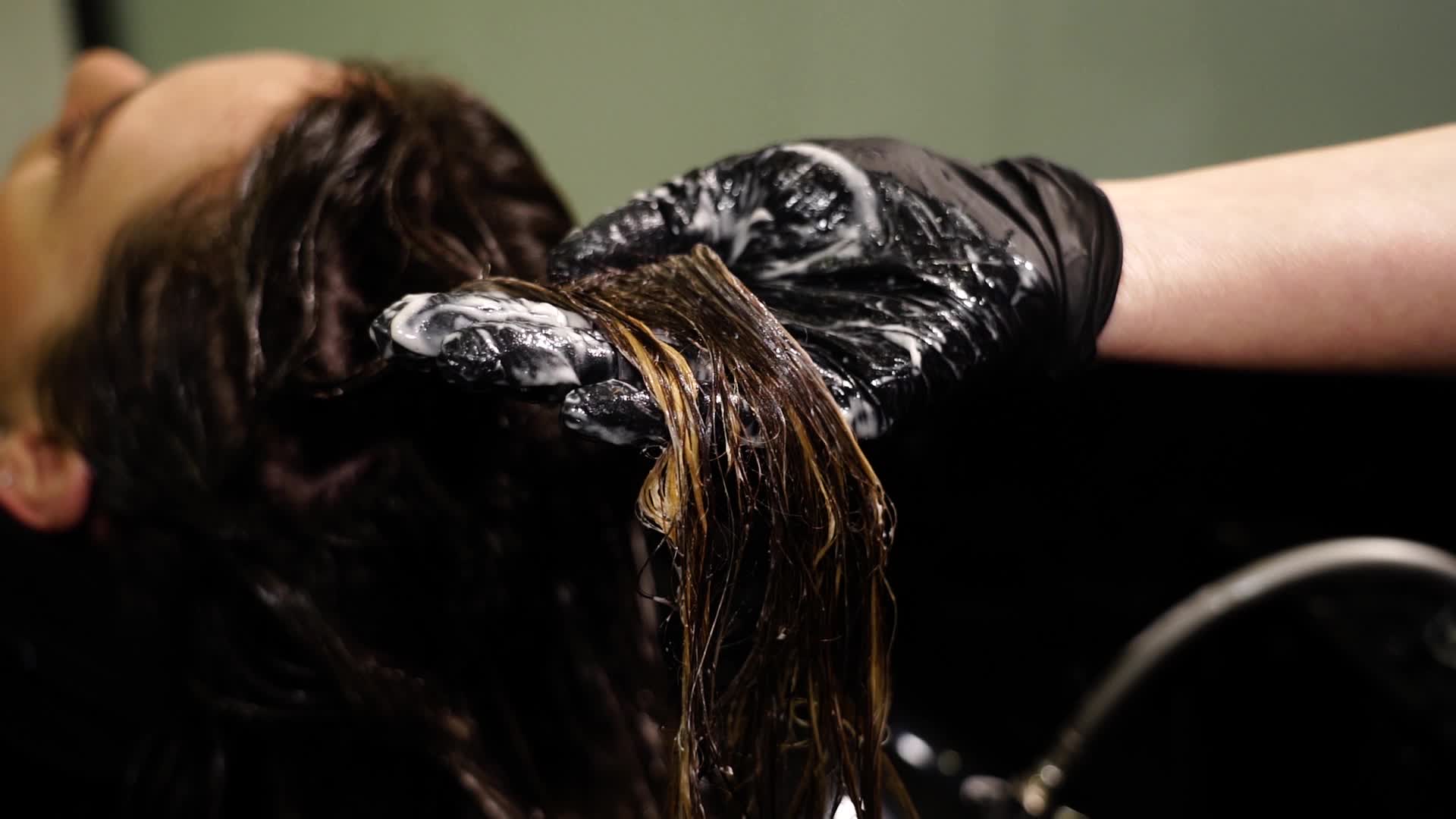 Hairstylist in glove pours lotion on long lady hair in salon