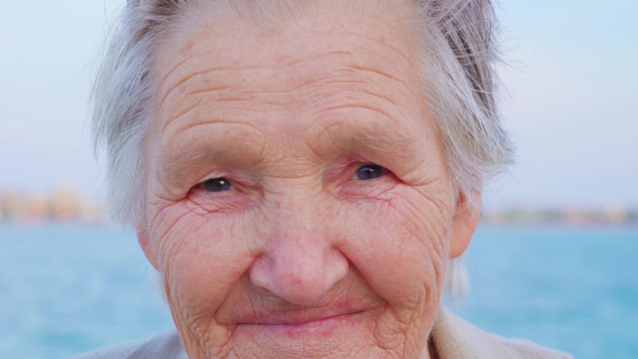 Senior grey-haired woman traveler with wrinkled face and smiling eyes looks into camera standing at lagoon in Chioggia extreme closeup