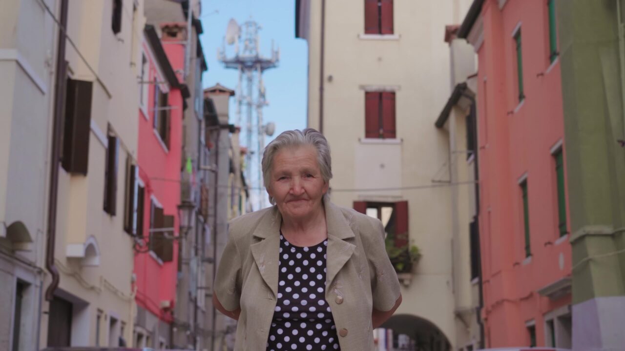 Elderly grey haired stooped woman traveler walks along street past historical colorful buildings in Chioggia smiling widely