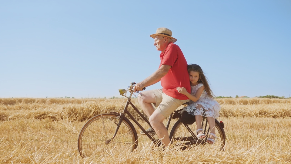 Little girl holds on to grandpa riding bicycle in field