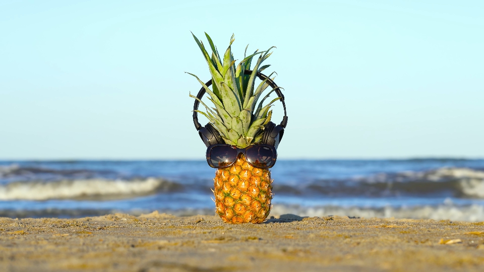 Pineapple with glasses and headphones on the sea sand